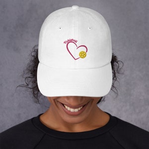May include: White baseball cap with a curved brim. The cap features an embroidered design of a heart and a pickleball, with the word "Pickleball" above the heart. The pickleball is yellow, and the heart and text are a deep pink.