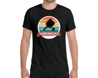 May include: A dark heather grey t-shirt featuring a vintage-style graphic of a pickleball paddle and ball. The graphic is set against a sunset background with orange, yellow, red, and blue stripes. The text "Pickleball" is written in a retro font below the graphic.