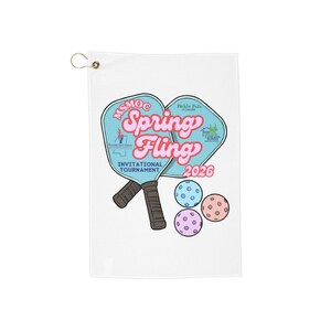May include: White towel with a design featuring two pickleball paddles and three balls. The design includes the text "Spring Fling Invitational Tournament 2026" in pink and blue. A gold grommet is at the top.