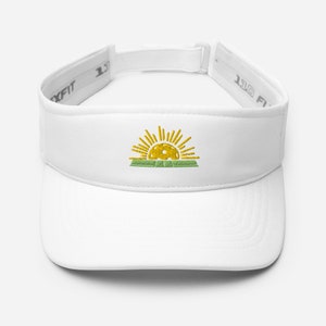 May include: White visor with a yellow sun and green grass embroidered on the front. The visor has a white adjustable strap with the text "1 SIZE FITS ALL" on both sides.