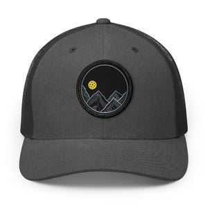 May include: A charcoal grey trucker hat featuring a circular patch on the front. The patch displays a black background with white line-art mountain peaks and a yellow pickleball positioned in the sky to represent a moon above the mountains.