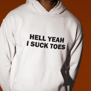 Op de afbeelding: Witte hoodie met de zwarte tekst "HELL YEAH I SUCK TOES". De sweatshirt heeft een voorzak en een capuchon met trekkoord. De achtergrond is effen oranje.