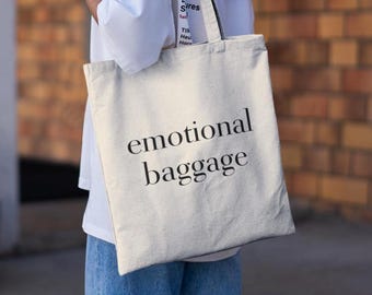 Puede incluir: Una persona lleva una bolsa de lona blanca con el texto "emotional baggage" impreso en negro.