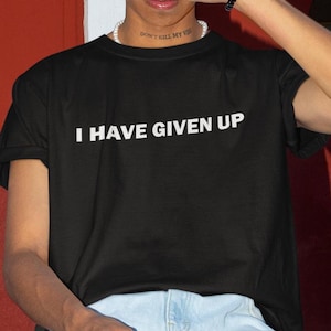 Può includere: Una maglietta nera con la scritta "I HAVE GIVEN UP" stampata in bianco sul davanti. La maglietta è indossata da una persona con una collana che dice "DON'T KILL MY VIBE".
