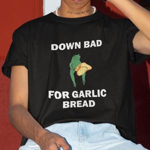 Könnte beinhalten: Schwarzes T-Shirt mit weißem Text, der "Down Bad for Garlic Bread" lautet. Ein grüner Frosch hält ein Stück Brot.