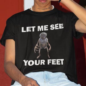 Può includere: Maglietta nera con il testo "Let me see your feet" in bianco. Sotto il testo è raffigurata una creatura con arti lunghi e un viso rugoso.