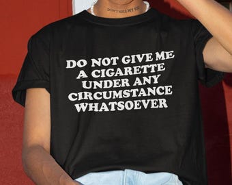 Könnte beinhalten: Schwarzes T-Shirt mit dem weißen Text "DO NOT GIVE ME A CIGARETTE UNDER ANY CIRCUMSTANCE WHATSOEVER". Das Shirt hat kurze Ärmel und einen Rundhalsausschnitt. Die Person trägt eine Perlenkette und hellblaue Jeans.