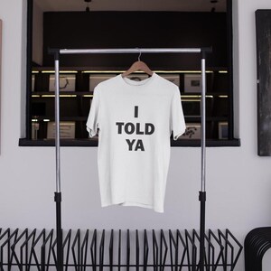 May include: White t-shirt with black text that reads "I TOLD YA" hanging on a clothing rack.