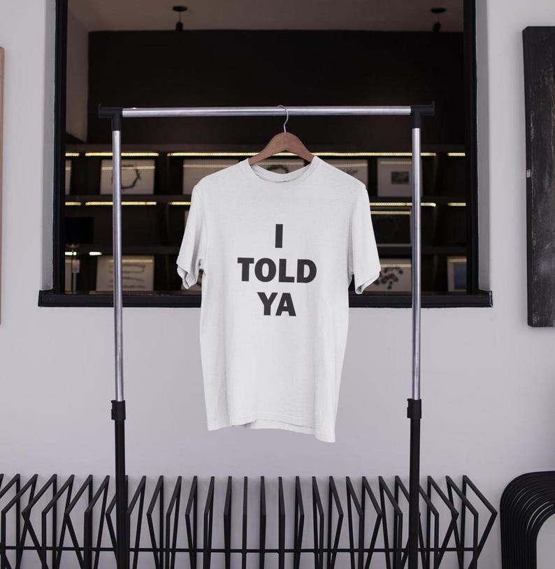 May include: White t-shirt with black text that reads "I TOLD YA" hanging on a clothing rack.