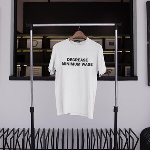 May include: White t-shirt with the text "DECREASE MINIMUM WAGE" printed in black on the front. The shirt is hanging on a black metal rack.