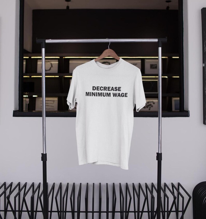 May include: White t-shirt with the text "DECREASE MINIMUM WAGE" printed in black on the front. The shirt is hanging on a black metal rack.