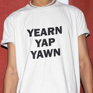 May include: White t-shirt featuring the words "YEARN YAP YAWN" in bold black text. The shirt has a crew neck and short sleeves. The image shows the upper body of a person wearing the t-shirt.