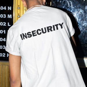 Könnte beinhalten: Weißes T-Shirt mit dem Wort "INSECURITY" in Schwarz auf dem Rücken.