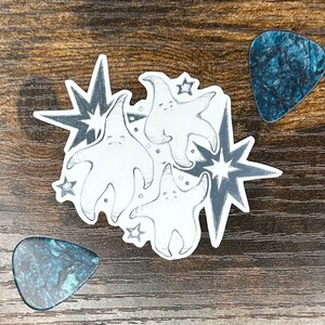May include: A sticker featuring three cartoon starfish with smiling faces, surrounded by starbursts and small stars. Two blue guitar plectrums are placed on a wooden surface near the sticker.