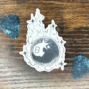 May include: A black and white sticker with an eight ball design in a circular frame, with flames and stars. Two guitar picks with a blue and black marbled pattern are on either side of the sticker. The items are on a wooden surface.