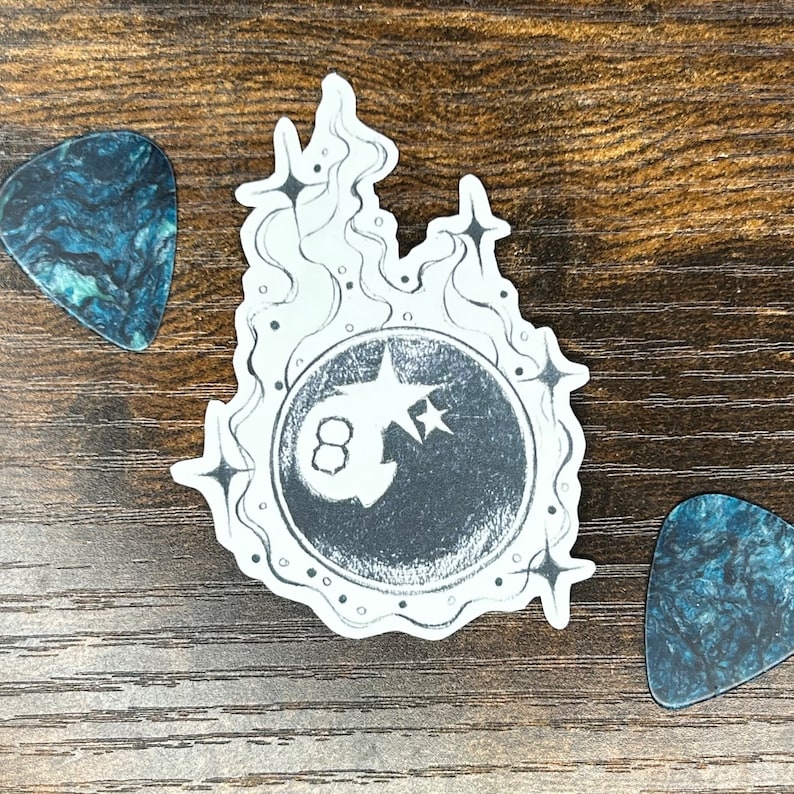 May include: A black and white sticker with an eight ball design in a circular frame, with flames and stars. Two guitar picks with a blue and black marbled pattern are on either side of the sticker. The items are on a wooden surface.