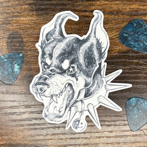 May include: A monochrome sticker depicting an aggressive dog with a spiked collar, baring its teeth. The illustration has a detailed, hand-drawn style. The sticker is edged in white and is set against a wooden background with guitar picks.