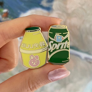 Könnte beinhalten: Zwei goldfarbene Emaille-Pins, einer in Form eines Milchkartons mit dem koreanischen Text "바나나맛 우유" und einem Comic-Kaninchen, der andere in Form einer Sprite-Dose mit einer blauen Comicfigur.