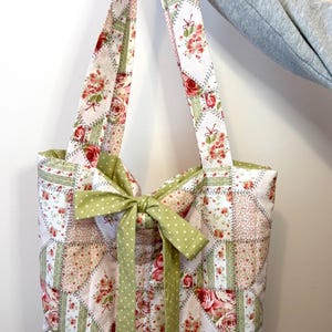 May include: A patchwork tote bag with floral and green accents. The bag features a repeating pattern of squares with red roses and green details. A green polka dot bow adorns the front, and the straps are made of the same floral fabric.
