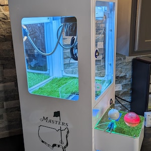 May include: White and green golf-themed claw machine with a Masters logo. The machine has a green roof and a display area with artificial turf. A small drawer holds golf balls and a miniature golfer.