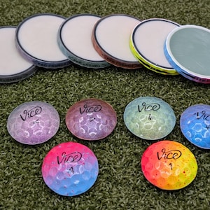 May include: An assortment of golf balls and ball markers on a green surface. The golf balls are multi-colored with the word "Vice" printed on them. The ball markers are white with colored rims and the text "BAD COSMOS".
