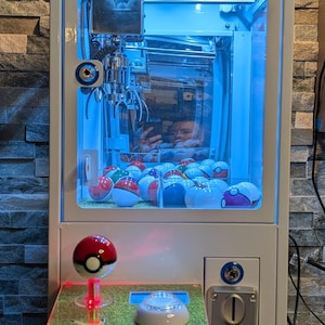 May include: A white Pokemon-themed claw machine with a blue roof and a glass front. The machine is filled with colorful balls and has a red and white Pokeball on the bottom. The machine has a coin slot and a button to operate the claw.