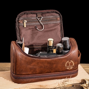 Op de afbeelding: Bruine leren reistas voor toiletartikelen met een ritssluiting bovenaan en een apart compartiment. De tas heeft een haak om op te hangen en een gepersonaliseerde inscriptie met de tekst "T Timothy".