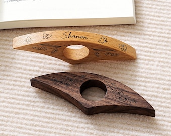May include: Two wooden thumb page holders. One is light-colored with butterfly and floral engravings, and the name "Shanon". The other is dark brown with floral engravings. Both have a circular opening for the thumb.