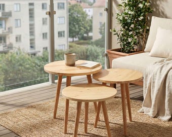 May include: A set of three round, light-colored wooden nesting tables with tapered legs. The tables are arranged in a cluster on a textured woven rug. A ceramic mug and a small book are placed on the surface of the largest table.