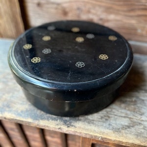 Puede incluir: Una caja redonda negra con una tapa decorada con diseños de flores doradas y plateadas. La caja está hecha de madera lacada y tiene un acabado liso y brillante.