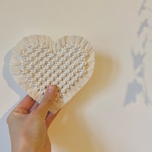 Peut inclure: Une suspension murale en macramé en forme de cœur blanche avec un bord frangé.