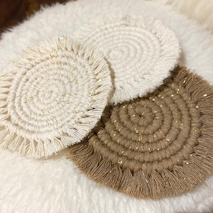Peut inclure: Trois dessous-de-verre ronds en macramé avec des franges. Deux sont blancs avec des accents dorés et un est brun avec des accents dorés.