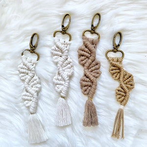 May include: Four macrame keyrings in neutral shades: off-white, cream, tan, and gold. Each keyring has a spiral design, a tassel, and a bronze-coloured clasp. The keyrings are displayed on a fluffy, white surface.
