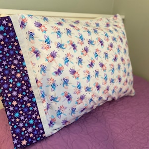 May include: A white pillowcase with a purple and blue unicorn pattern. The pillowcase has a purple border with a white and blue star pattern.