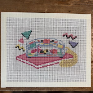 May include: A needlepoint canvas featuring a clear, multi-compartment desk organizer sitting on a pink rectangular mat. The design includes colorful geometric shapes like triangles and zigzags in teal, pink, yellow, and purple surrounding the organizer.
