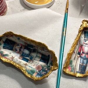 Puede incluir: Dos conchas de ostra decoradas con un borde de pintura metálica dorada. Los interiores presentan un diseño navideño con un soldado cascanueces. Un pincel turquesa se encuentra entre las conchas, sugiriendo una artesanía.
