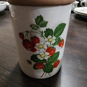 May include: A cylindrical white ceramic canister featuring a botanical illustration of red strawberries, green leaves, and white flowers. The container is topped with a smooth, round, light-colored wooden lid that sits flush on the top rim.
