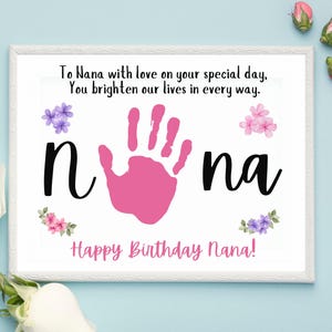 Può includere: Una stampa incorniciata con il testo "To Nana with love on your special day, You brighten our lives in every way." La parola "Nana" è in grandi lettere nere con un'impronta di mano rosa al centro. Le parole "Happy Birthday Nana!" sono in basso.