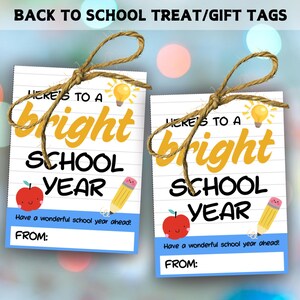 Puede incluir: Dos etiquetas de regalo rectangulares con el texto "Here's to a bright school year" en amarillo y negro. Cada etiqueta presenta una manzana roja, un lápiz amarillo y una bombilla. Las etiquetas están atadas con hilo.