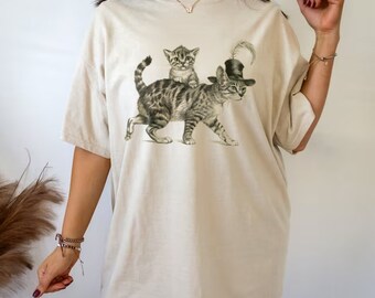 Könnte beinhalten: Ein beigefarbenes Oversize-T-Shirt mit einem grafischen Aufdruck von zwei Katzen. Die größere Katze trägt einen schwarzen Hut mit einer Feder. Die kleinere Katze sitzt auf dem Rücken der größeren Katze.