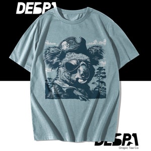 May include: A light blue t-shirt with a graphic print of a koala wearing a pirate hat and an eye patch. The koala is surrounded by trees and clouds.