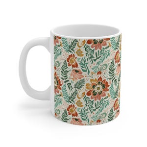 May include: White ceramic mug with a floral pattern. The design features red, orange, and pink flowers with green leaves and accents on a cream background. The mug has a curved handle, perfect for tea or coffee.