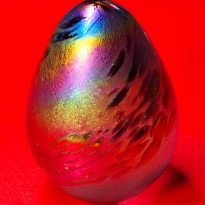May include: A colorful, iridescent glass egg-shaped sculpture. The egg features a blend of colors, including blues, greens, yellows, pinks, and reds. The surface has a textured, organic pattern. The sculpture is set against a bright red background.