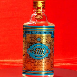 May include: A clear glass bottle of 4711 Original Eau de Cologne. The label features gold filigree and turquoise accents, with the number 4711 prominently displayed. The bottle has a gold cap and is set against a red background.