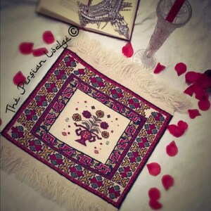 May include: A white square rug with a red, yellow, and blue geometric pattern and a floral design in the center. The rug has fringe around the edges and is surrounded by rose petals.