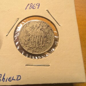 May include: A silver coin from 1869, displayed in a protective holder. The coin features a shield design and is encased in a cardboard frame. The year "1869" is handwritten in blue ink above the coin.