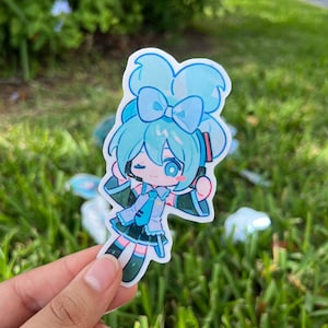 K&ouml;nnte beinhalten: Ein blauer und gr&uuml;ner Anime-Charakter-Aufkleber mit einer blauen Schleife und Kopfh&ouml;rern. Der Charakter ist ein junges M&auml;dchen mit einer leuchtend blauen Schleife und einem hellblauen und gr&uuml;nen Outfit.