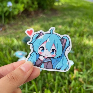 K&ouml;nnte beinhalten: Ein Aufkleber mit einem blauen und schwarzen Anime-Charakter mit einem rosa Herzen &uuml;ber ihrem Kopf. Der Aufkleber befindet sich auf einem gr&uuml;nen grasbewachsenen Hintergrund.