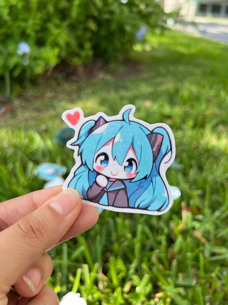 K&ouml;nnte beinhalten: Ein Aufkleber mit einem blauen und schwarzen Anime-Charakter mit einem rosa Herzen &uuml;ber ihrem Kopf. Der Aufkleber befindet sich auf einem gr&uuml;nen grasbewachsenen Hintergrund.