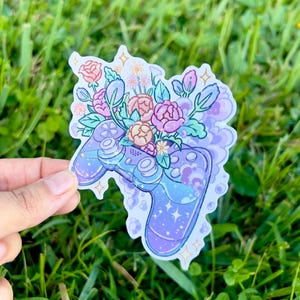 Puede incluir: Una pegatina con un mando de videojuegos adornado con un ramo floral. El mando tiene un degradado púrpura y azul con detalles de estrellas. Las flores son rosas, melocotón y blancas con hojas verdes.
